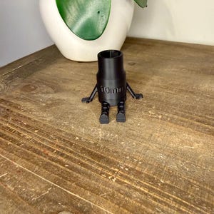 Peut inclure: Une douille noire de 10 mm avec des bras et des pieds attach&eacute;s, ressemblant &agrave; un petit robot. La douille est pos&eacute;e sur une surface en bois, avec un pot blanc et une plante verte en arri&egrave;re-plan.