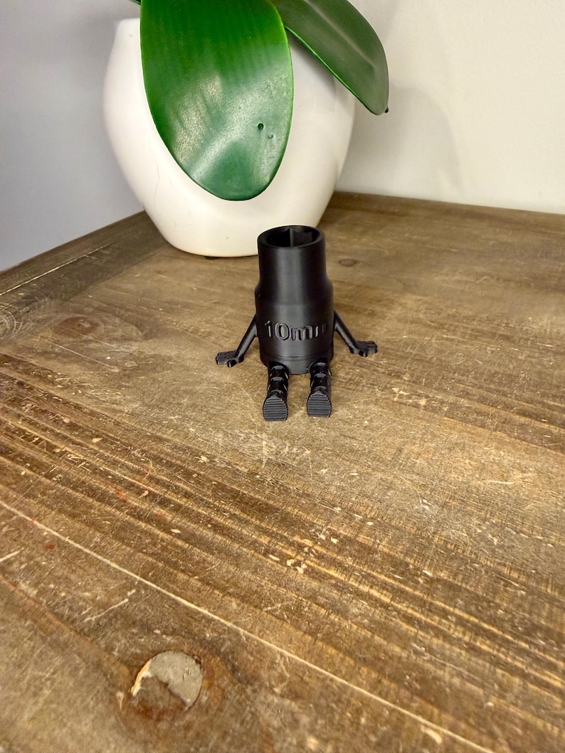 Peut inclure: Une douille noire de 10 mm avec des bras et des pieds attach&eacute;s, ressemblant &agrave; un petit robot. La douille est pos&eacute;e sur une surface en bois, avec un pot blanc et une plante verte en arri&egrave;re-plan.