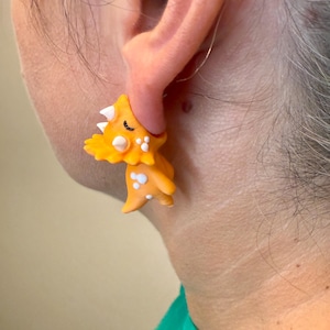 May include: Orange dinosaur earrings with white accents and black eyes. The earrings are a fun, whimsical design, perfect for anyone who loves dinosaurs or unique jewelry.