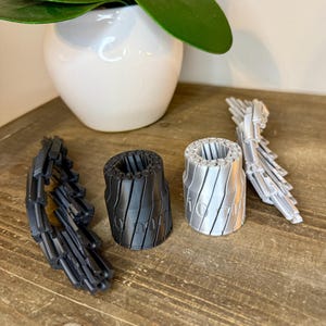 May include: A collection of 3D-printed objects in black and silver, including two cylindrical pieces with vertical grooves and the text "10/10" on one. Also, there are two articulated, segmented pieces. A white vase with green leaves is in the background.