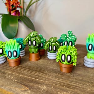 K&ouml;nnte beinhalten: Eine Sammlung von neun handbemalten Keramikfiguren verschiedener Pflanzen, jede mit einem einzigartigen gr&uuml;n-wei&szlig;en Farbschema und gro&szlig;en, schwarzen Augen. Die Figuren sind auf einer Holzoberfl&auml;che angeordnet.