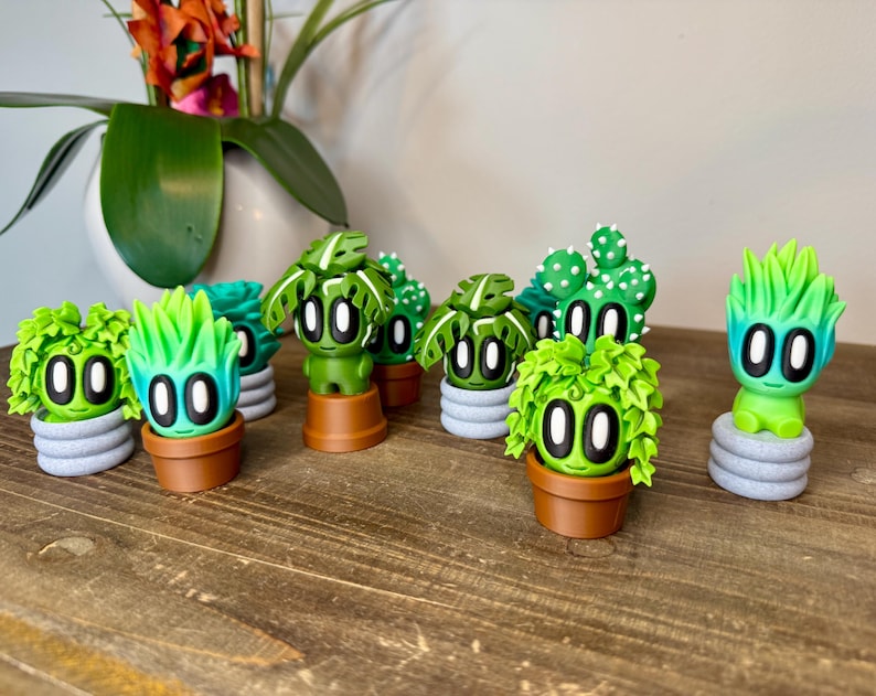 K&ouml;nnte beinhalten: Eine Sammlung von neun handbemalten Keramikfiguren verschiedener Pflanzen, jede mit einem einzigartigen gr&uuml;n-wei&szlig;en Farbschema und gro&szlig;en, schwarzen Augen. Die Figuren sind auf einer Holzoberfl&auml;che angeordnet.