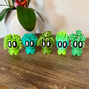 K&ouml;nnte beinhalten: F&uuml;nf kleine gr&uuml;ne Figuren mit verschiedenen pflanzenartigen Frisuren. Jede Figur hat zwei gro&szlig;e schwarze Augen und einen kleinen wei&szlig;en Mund. Die Figuren sind in einer Reihe auf einer Holzoberfl&auml;che angeordnet.