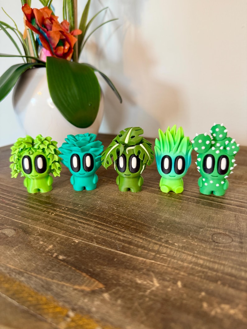 K&ouml;nnte beinhalten: F&uuml;nf kleine gr&uuml;ne Figuren mit verschiedenen pflanzenartigen Frisuren. Jede Figur hat zwei gro&szlig;e schwarze Augen und einen kleinen wei&szlig;en Mund. Die Figuren sind in einer Reihe auf einer Holzoberfl&auml;che angeordnet.