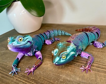 May include: Two iridescent lizard sculptures in shades of teal, purple, and bronze rest on a wooden surface. The lizards have detailed features, including yellow eyes and articulated limbs. A white ceramic pot with a green plant is in the background.