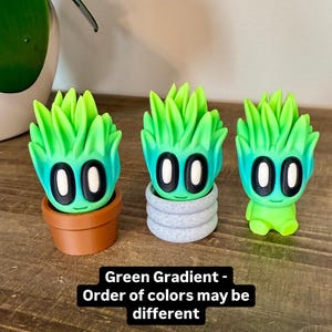 K&ouml;nnte beinhalten: Drei kleine Figuren in gr&uuml;nem Farbverlauf mit schwarzen Augen und einem l&auml;chelnden Mund. Die Figuren sind in verschiedenen Posen, eine ist in einem braunen Topf, eine in einem grauen Topf und eine steht alleine. Der Text "Green Gradient - Order of colors may be different" befindet sich unter den Figuren.