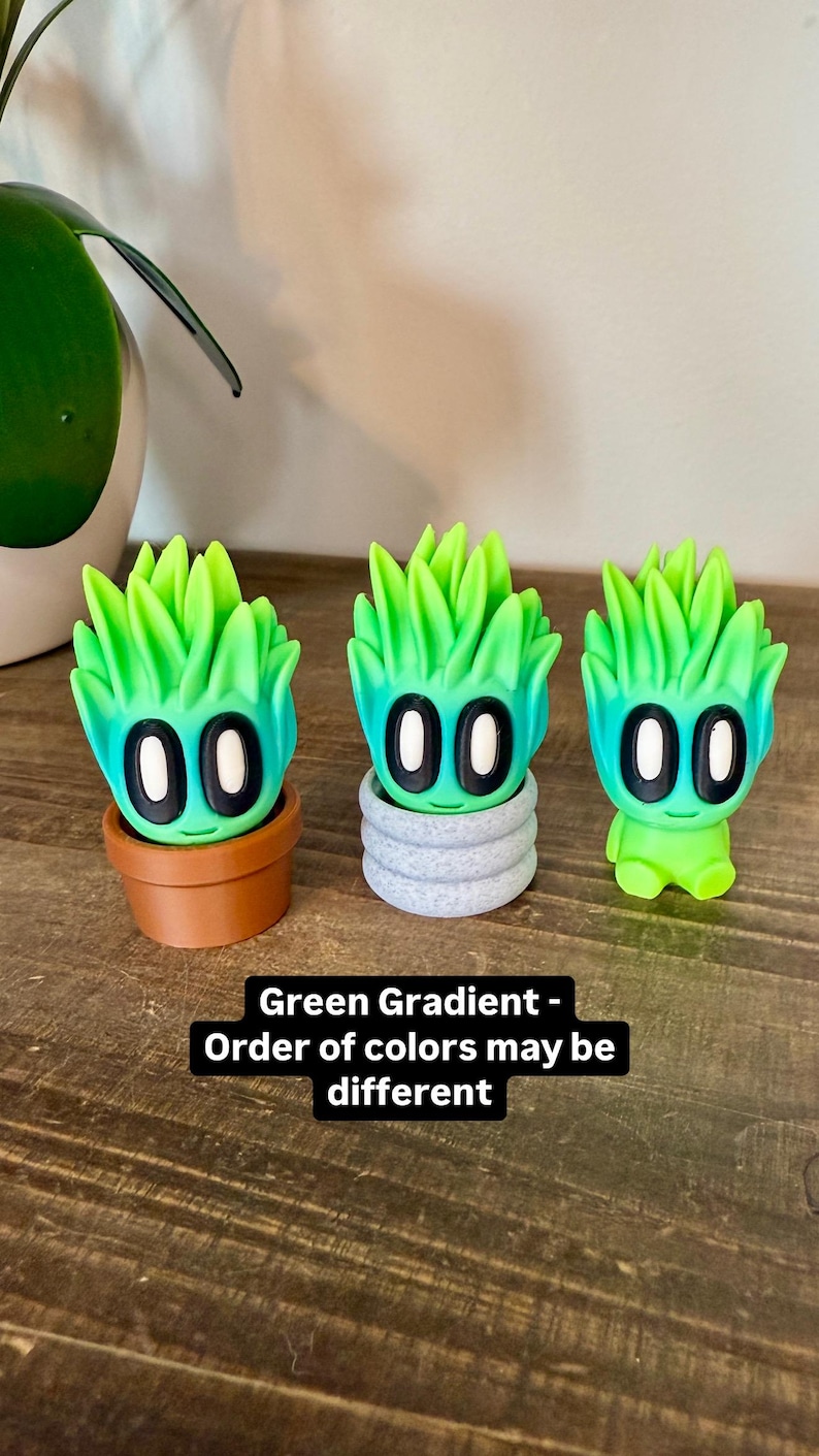 K&ouml;nnte beinhalten: Drei kleine Figuren in gr&uuml;nem Farbverlauf mit schwarzen Augen und einem l&auml;chelnden Mund. Die Figuren sind in verschiedenen Posen, eine ist in einem braunen Topf, eine in einem grauen Topf und eine steht alleine. Der Text "Green Gradient - Order of colors may be different" befindet sich unter den Figuren.
