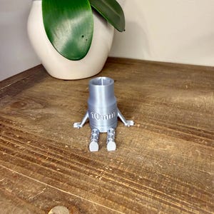 Peut inclure: Une cl&eacute; &agrave; douille argent&eacute;e imprim&eacute;e en 3D, avec des bras et des jambes, et le texte "10mm" imprim&eacute; sur le c&ocirc;t&eacute;. La cl&eacute; est pos&eacute;e sur une surface en bois, avec un pot blanc et une plante verte en arri&egrave;re-plan.