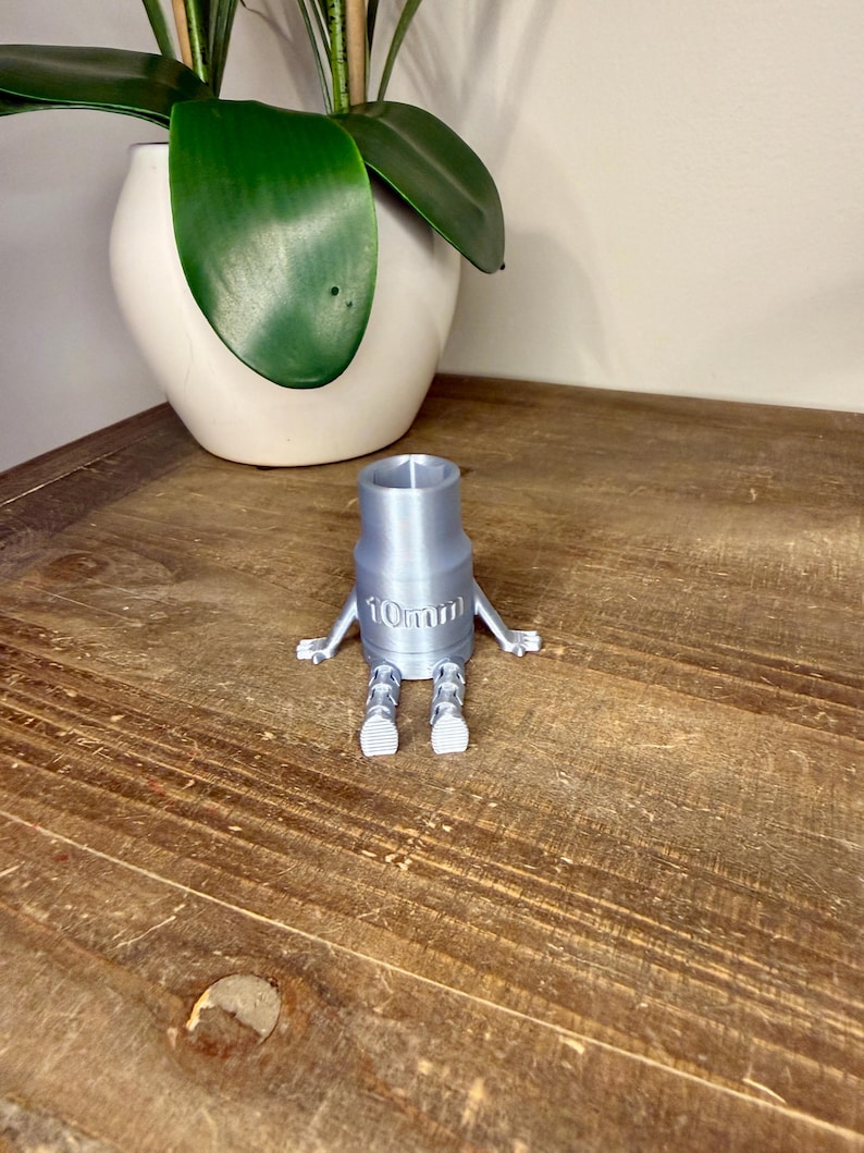 Peut inclure: Une cl&eacute; &agrave; douille argent&eacute;e imprim&eacute;e en 3D, avec des bras et des jambes, et le texte "10mm" imprim&eacute; sur le c&ocirc;t&eacute;. La cl&eacute; est pos&eacute;e sur une surface en bois, avec un pot blanc et une plante verte en arri&egrave;re-plan.
