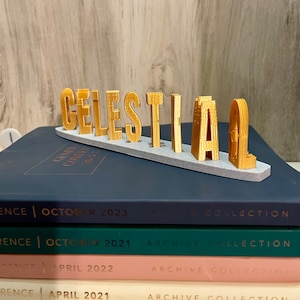 May include: Gold-colored letters spelling out the word "CELESTIAL" on a gray base. The letters are 3D printed and have a textured surface.