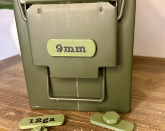 May include: An olive green metal ammo box with a hinged lid and a handle. The box has a label that reads "9mm". Several olive green accessories with the text "12ga" are in front of the box. The items are on a wooden surface.