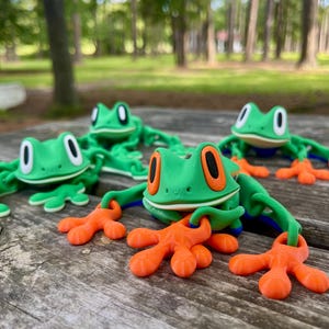 May include: Four green frog figurines with orange feet and blue eyes. The frogs are posed in a sitting position with their legs outstretched.