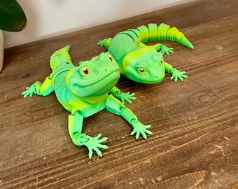 May include: Two green and yellow lizard figurines are displayed on a wooden surface. The lizards have detailed features, including eyes, legs, and segmented tails. The color gradient adds depth to the design, creating a playful and decorative item.