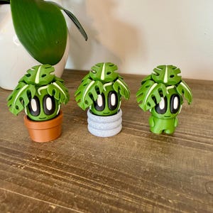 K&ouml;nnte beinhalten: Drei kleine gr&uuml;ne Monstera-Pflanzenfiguren mit wei&szlig;en Akzenten, jede in einem anderen Topf. Die T&ouml;pfe sind braun, grau und gr&uuml;n.