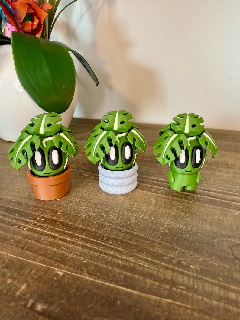 K&ouml;nnte beinhalten: Drei kleine gr&uuml;ne Monstera-Pflanzenfiguren mit wei&szlig;en Akzenten, jede in einem anderen Topf. Die T&ouml;pfe sind braun, grau und gr&uuml;n.