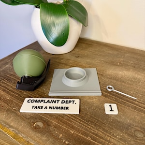 May include: A collection of objects including a green sphere, a black clip, a grey square with a circular aperture, a small metal tool, and a white sign reading "COMPLAINT DEPT. TAKE A NUMBER". A white tile with the number "1" is also present.