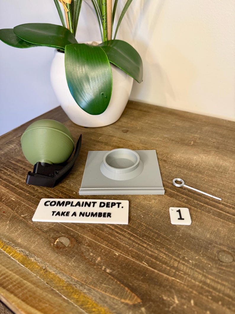 May include: A collection of objects including a green sphere, a black clip, a grey square with a circular aperture, a small metal tool, and a white sign reading "COMPLAINT DEPT. TAKE A NUMBER". A white tile with the number "1" is also present.