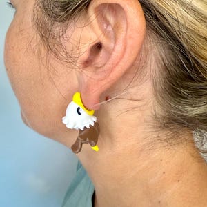 May include: A pair of novelty earrings shaped like cartoon eagles. The earrings feature a white head, brown body, and yellow beak and feet. The earrings are attached to a post.