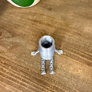 Peut inclure: Une cl&eacute; &agrave; douille grise claire imprim&eacute;e en 3D avec des bras et des jambes, ressemblant &agrave; un robot. La cl&eacute; a une ouverture hexagonale et se trouve sur une surface en bois. Un pot blanc et vert est en arri&egrave;re-plan.