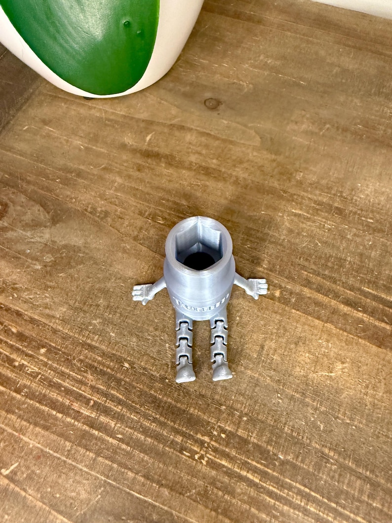 Peut inclure: Une cl&eacute; &agrave; douille grise claire imprim&eacute;e en 3D avec des bras et des jambes, ressemblant &agrave; un robot. La cl&eacute; a une ouverture hexagonale et se trouve sur une surface en bois. Un pot blanc et vert est en arri&egrave;re-plan.