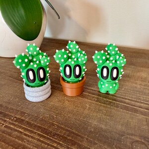 K&ouml;nnte beinhalten: Drei gr&uuml;ne Kaktus-Figuren mit wei&szlig;en Punkten und schwarzen Augen. Der Kaktus links steht in einem grauen Topf, der mittlere Kaktus in einem braunen Topf und der Kaktus rechts steht alleine.