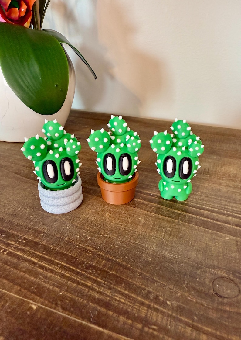 K&ouml;nnte beinhalten: Drei gr&uuml;ne Kaktus-Figuren mit wei&szlig;en Punkten und schwarzen Augen. Der Kaktus links steht in einem grauen Topf, der mittlere Kaktus in einem braunen Topf und der Kaktus rechts steht alleine.