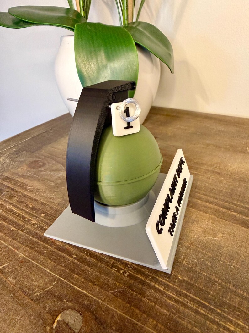 May include: A novelty desk ornament shaped like a green grenade with a black handle and a white tag with the number 1. A white sign reads "COMPLAINT DEPT. TAKE A NUMBER."