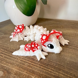 May include: A white, whimsical dragon figurine with red and white mushroom accents. The dragon has a detailed, textured body and black eyes. The mushrooms feature red caps with white polka dots. The figurine is displayed on a wooden surface.