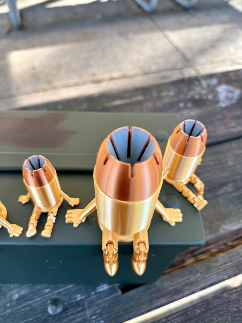 May include: Four gold-coloured, 3D printed figurines shaped like bullets with articulated legs and arms. The figurines are sitting on a green surface.