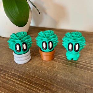 K&ouml;nnte beinhalten: Drei gr&uuml;ne Sukkulenten-Figuren mit wei&szlig;en Augen und einem l&auml;chelnden Mund. Die Sukkulenten stehen in verschiedenen T&ouml;pfen, einer in einem grauen Topf, einer in einem braunen Topf und einer steht alleine.