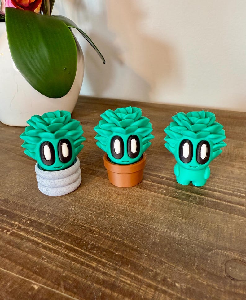 K&ouml;nnte beinhalten: Drei gr&uuml;ne Sukkulenten-Figuren mit wei&szlig;en Augen und einem l&auml;chelnden Mund. Die Sukkulenten stehen in verschiedenen T&ouml;pfen, einer in einem grauen Topf, einer in einem braunen Topf und einer steht alleine.