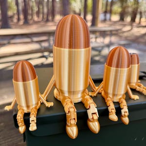 May include: Three gold-coloured bullet-shaped figurines with articulated legs are sitting on a green metal ammo can. The figurines are different sizes.