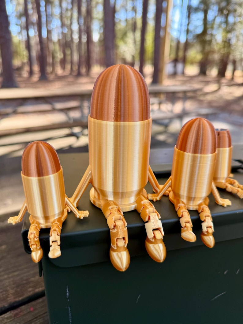 May include: Three gold-coloured bullet-shaped figurines with articulated legs are sitting on a green metal ammo can. The figurines are different sizes.
