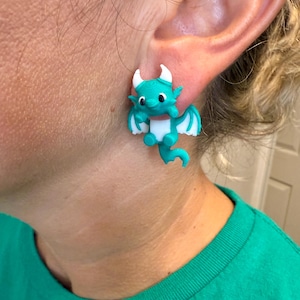 May include: A pair of teal dragon earrings. Each earring features a small, stylized dragon with white accents on the wings, belly, and horns. The earrings are designed to be worn in the ear, showcasing a whimsical and fantasy-themed accessory.