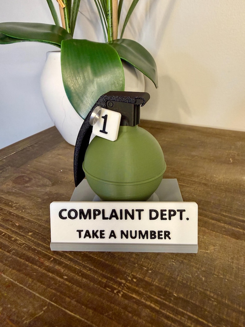 May include: A novelty desk ornament resembling a green hand grenade, featuring a black handle and a white tag with the number 1. The sign below says "COMPLAINT DEPT. TAKE A NUMBER."