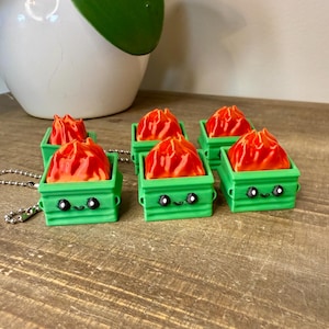 May include: Six green miniature trash cans with cartoon faces, each containing a red and orange flame. The trash cans are on a wooden surface, with a silver chain attached to one. A white pot with a green plant is in the background.