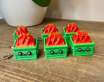 May include: Six green miniature trash cans with cartoon faces, each containing a red and orange flame. The trash cans are on a wooden surface, with a silver chain attached to one. A white pot with a green plant is in the background.