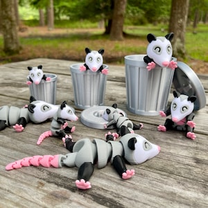 May include: A set of six 3D printed possum figurines. Five of the possums are in a trash can pose, while one is lying on its back with its legs in the air. The possums are all gray and white with pink accents.