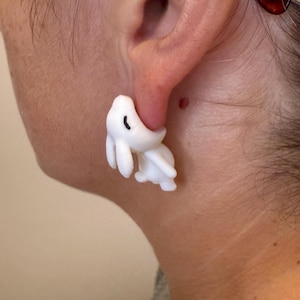 May include: A white bunny-shaped earring is worn on the ear. The earring has a curved body and two long ears. The bunny's eye is a small black dot. The earring is made of a smooth, solid material.