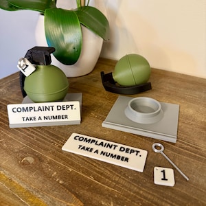 May include: A novelty desk set with a green grenade, a green sphere, and a grey base. The set includes a sign reading "COMPLAINT DEPT. TAKE A NUMBER" and a small number "1" tile. The items are displayed on a wooden surface.