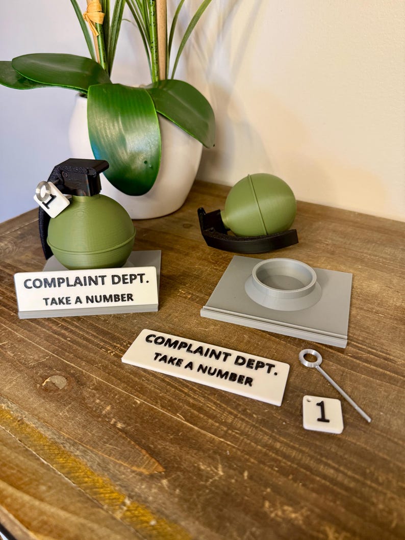 May include: A novelty desk set with a green grenade, a green sphere, and a grey base. The set includes a sign reading "COMPLAINT DEPT. TAKE A NUMBER" and a small number "1" tile. The items are displayed on a wooden surface.