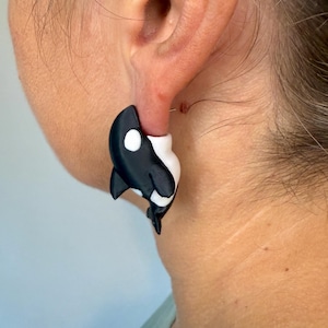 May include: Black and white orca earrings. The earrings are shaped like orcas, with a black body, white belly, and a white spot near the eye. The earrings are worn on the ear.