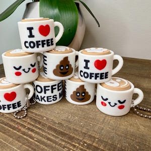May include: A set of seven white ceramic coffee mugs with different designs. The mugs feature a red heart with the text "I love coffee", a poop emoji, a yin yang symbol, and a smiley face.
