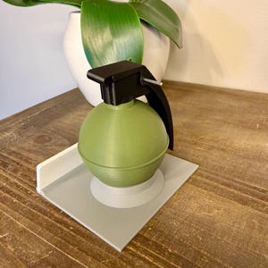 May include: A green, spherical decorative object with a black handle and top, resembling a grenade. It sits on a grey square base with a white border. The object is placed on a wooden surface.