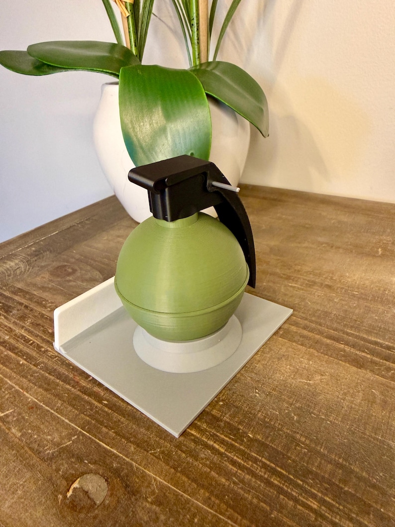 May include: A green, spherical decorative object with a black handle and top, resembling a grenade. It sits on a grey square base with a white border. The object is placed on a wooden surface.