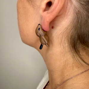 May include: A brown beaver-shaped earring with a black tail is worn on a person's ear. The earring is a small, detailed sculpture of the animal. The background is a neutral color, highlighting the earring's design.