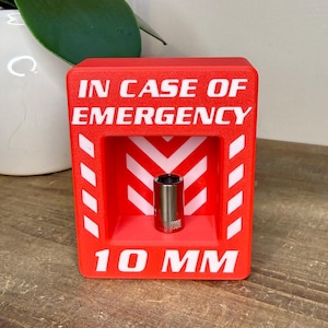 May include: A red box with the text "IN CASE OF EMERGENCY" and "10 MM" in white. Inside the box is a silver socket. The box has white chevron and stripe accents.
