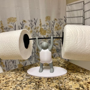 May include: A white toilet roll holder shaped like a person lifting weights. The holder has a black metal bar and two rolls of white toilet paper.