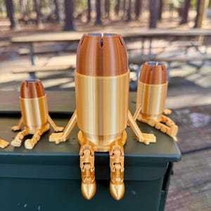 May include: Three gold-coloured bullet casings with articulated legs, sitting on a green metal ammo can. The casings are made of plastic and have a textured surface.