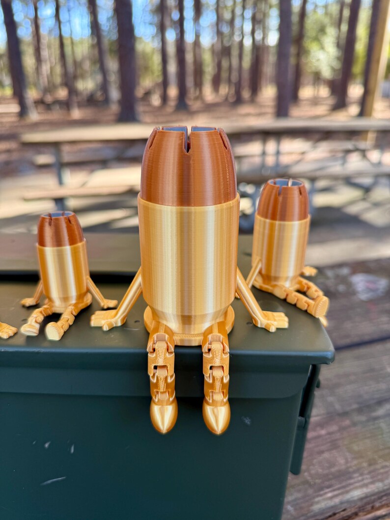 May include: Three gold-coloured bullet casings with articulated legs, sitting on a green metal ammo can. The casings are made of plastic and have a textured surface.